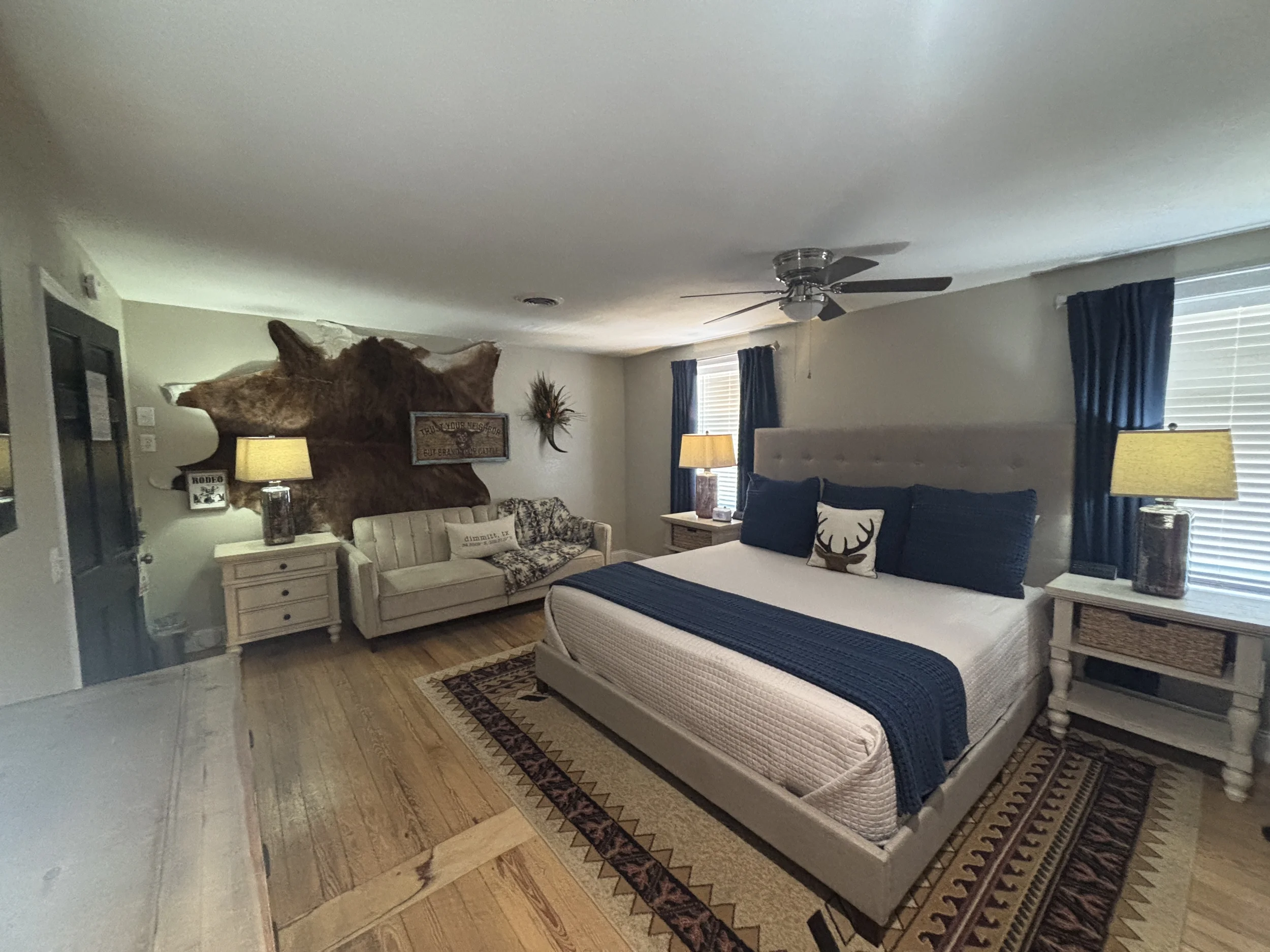 A spacious rustic-style bedroom featuring a large bed with a tufted beige headboard, white bedding, and navy blue pillows and throw. Matching white nightstands with lamps sit on each side of the bed. 