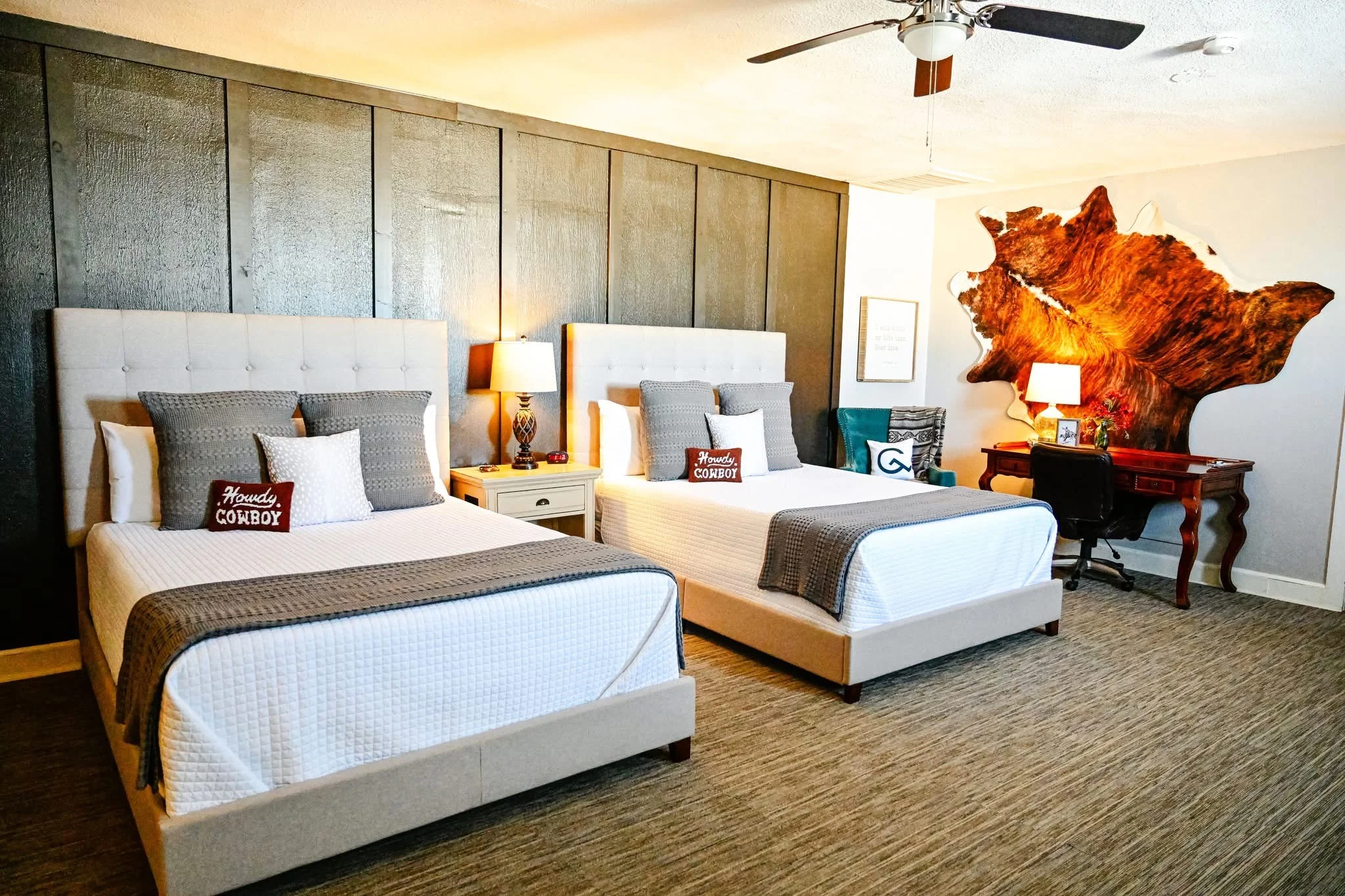 A stylish Western-themed guest room featuring two upholstered queen beds with tufted beige headboards, white bedding, and gray accent pillows. Each bed has a decorative “Howdy Cowboy” pillow. A shared